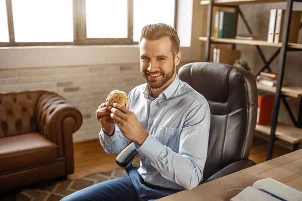 Jeune homme d'affaires beau assis sur une chaise et déjeuner dans son propre bureau. Il tient un hamburger et sourit à la caméra. Positif affamé jeune homme manger . — Photo