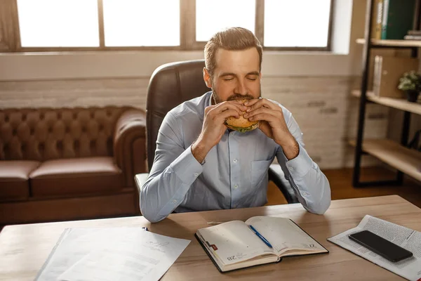 잘생긴 젊은 사업가 테이블과 물고 햄버거에 자신의 사무실에 앉아. 그는 점심 시간. 배고픈 젊은이 삼 킬 음식. — 스톡 사진