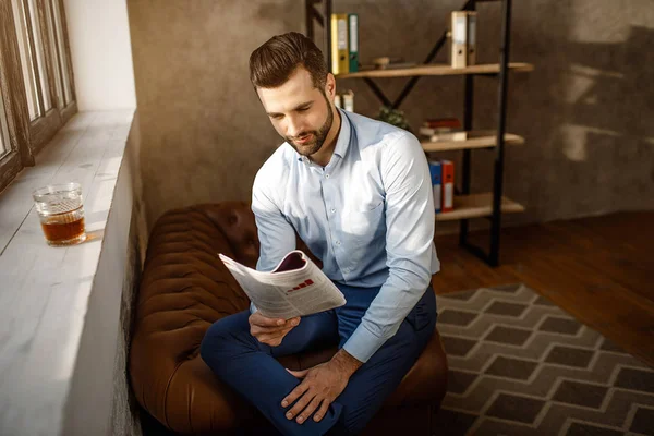 Young handsome businessman reading journal in his own office. He sit at window and read journal. Glass of whiskey stand on windowsill. Sunlight on wall.