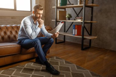 Genç yakışıklı düşünceli işadamı oturun ve kendi ofisinde günlüğü okuyun. O poz. Kendinden emin ve seksi. Gün ışığı.