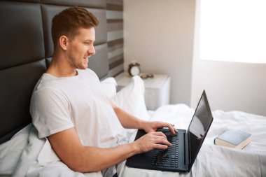 Yatak erken uyandığımız, neşeli delikanlıyı eser bulunmaktadır. Adam tutun laptop ve türü klavyede. Gülümseme ve olumlu. Gün ışığı.
