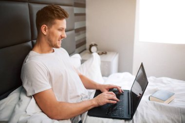 Cheerful nice young man work in bed early moring. Guy hold laptop and type on keyboard. Smile and positive. Daylight. clipart