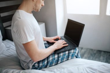 Genç adam bu sabah yatakta. O Pijama giyiyor. Adam klavye laptop yatak ve türü kenarında oturmak. O evde iş. Gün ışığı.