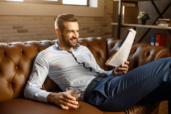 Joven hombre de negocios guapo sentarse en el sofá y leer en su propia oficina. Tiene un vaso de whisky en la mano y sonríe. Positivo y feliz. Bien parecido. . — Foto de Stock