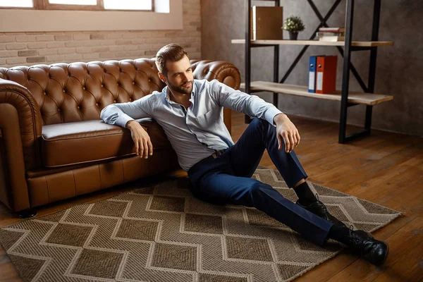 Jeune homme d'affaires beau assis sur le sol et pose dans son propre bureau. Il a l'air hétéro. Sexy jeune homme maigre au canapé . — Photo