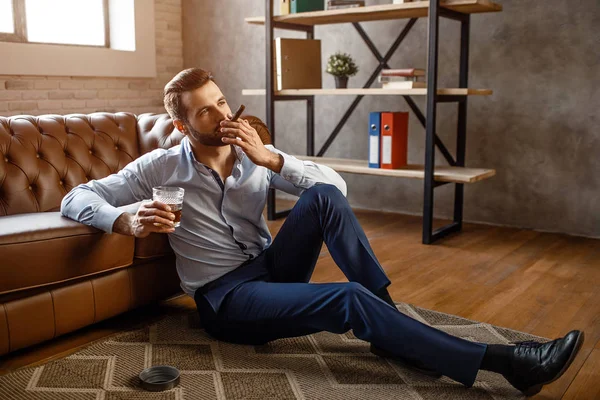 Jeune homme d'affaires beau fumer un cigare et tenir un verre de whisky dans son propre bureau. Il s'assoit par terre et a l'air confiant. Sexy jeune homme relaxant . — Photo