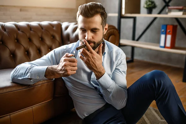 Young handsome businessman smoke cigar in his own office. He sit on floor and lean to sofa. Guy hold cigarette lighter. Sexy and concentrated.