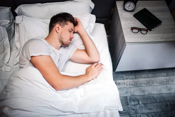Νεαρός άνδρας στο κρεβάτι το πρωί. Κάτοψη του κουρασμένος άνθρωπος στον ύπνο. Σκέπασε με την κουβέρτα λευκό. Το κεφάλι στο μαξιλάρι. — Φωτογραφία Αρχείου