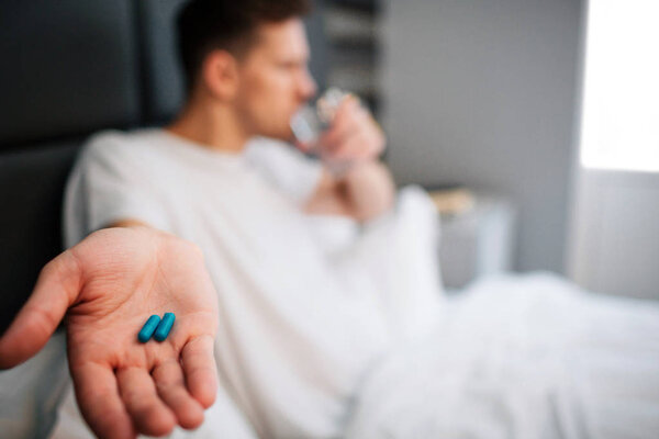 Молодой человек в постели рано утром. Он держит две капсулы на ладони. Пейте воду. Daylight
.