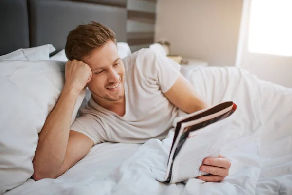 Cheerful nice young man lying in bed early morning. He read journal. Guy hold head on hand and smile. Positive nd relaxed. Daylight.