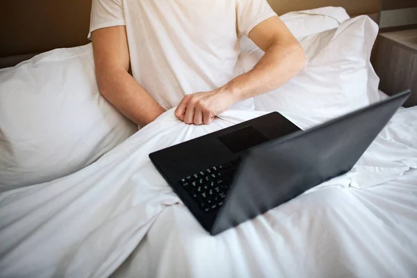 Genç adam sabah erkenden yatağa otur. O beyaz battaniye ve mastürbasyon altında elini tut. Bacakları üstünde laptop. Video izlerken.