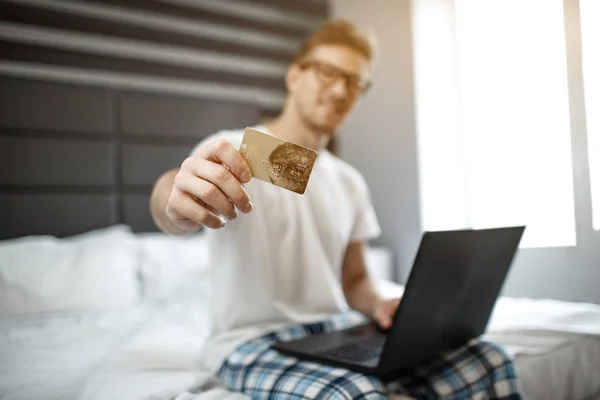 Nice young man sit on bed early morning. He show credit card on camera. Guy hold laptop. Electronic money.