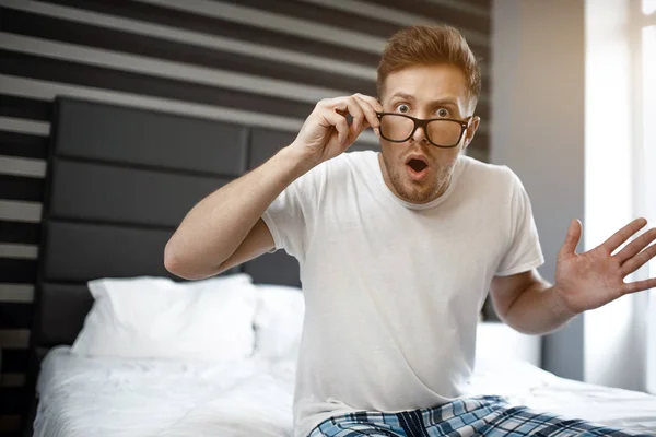 Zdumiony młody człowiek siedzieć na łóżku wcześnie rano. Spojrzeć na kamery i trzymać rękę na okulary. Przestraszony facet. — Zdjęcie stockowe