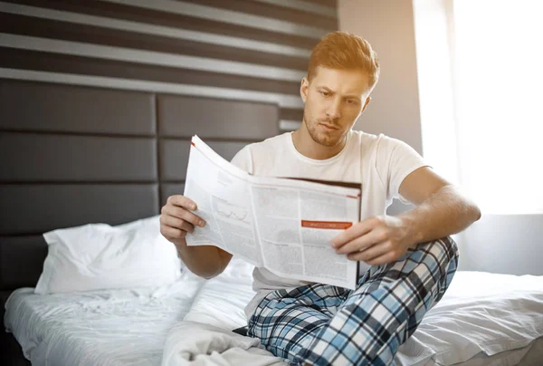 Serious thoughtful young man on bed early morning. He sit and read newspaper or journal. — Stock Photo, Image