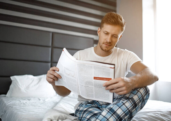 Young man on bed early morning. He sit and read journal. Small smile on face. Concentrated.