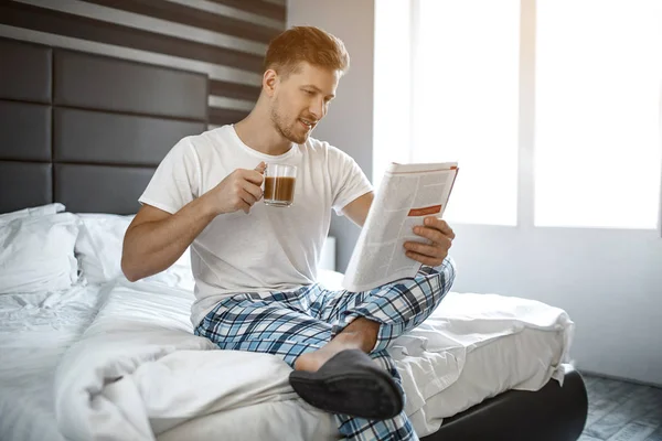 Młody człowiek na łóżku w nocy. Mu siedzieć i czytać czasopisma facet trzymać filiżankę kawy. Miły i wesoły. — Zdjęcie stockowe