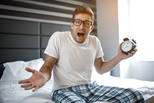 Young man on bed in morning. He overslept. Guy hold clock in hand and look on camera.