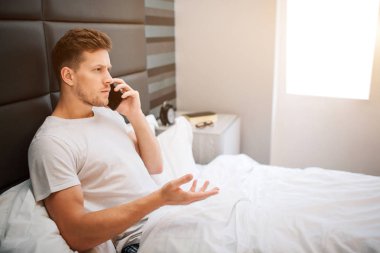 Genç adam bu sabah yatakta. Ciddi erkek model Oda oturmak ve telefon konuşmak. O beyaz battaniye ile örtülü. Gün ışığı. Sabah erkenden. Uyandı.