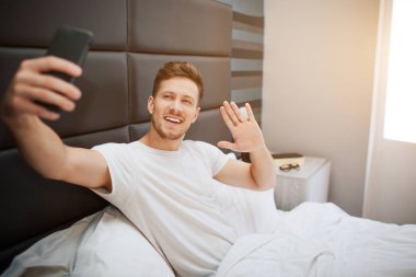 Bu sabah yatakta genç neşeli adam. O ve telefon kamera poz. Adam dalga el ve bir gülümseme ile. Gün ışığı.