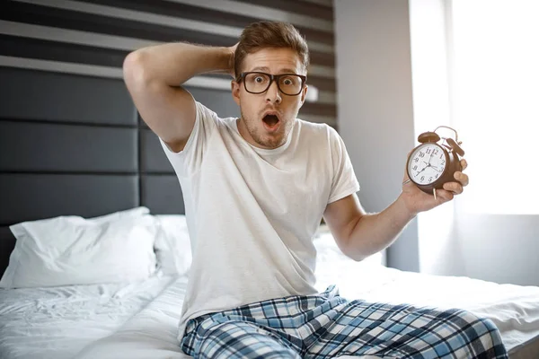Amazed and scarec young man on bed in morning. He look emotionally on camera. Guy hold clock in hand.