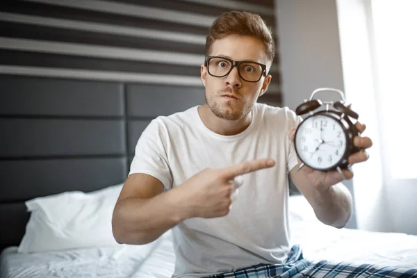 Ernstige angry young man op bed in de ochtend. Hij kijkt op camera en wijs op klok. — Stockfoto