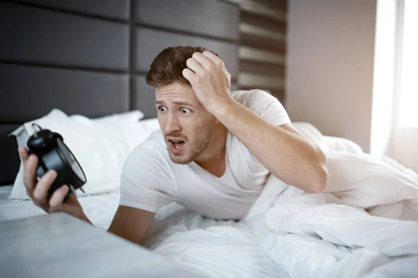 Emotional young man on bed in early morning. He overslept. Guy hold clock and look at it scared.