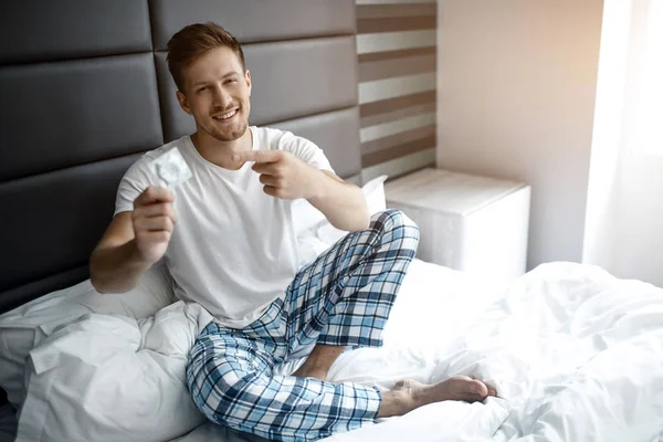 Young sexy man on bed early morning. He hold condom in hand and point on it. Smiling.