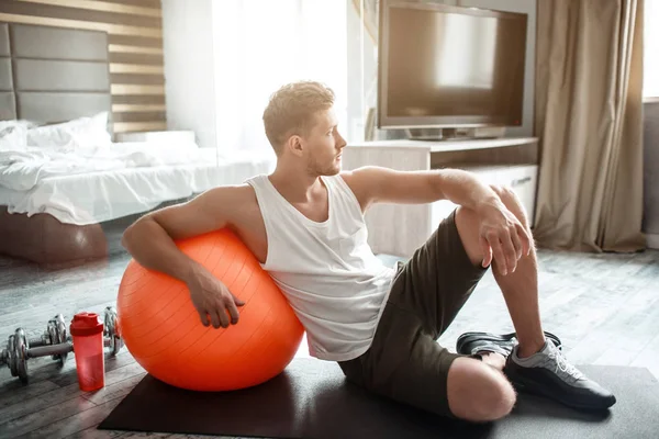 Der junge, gut gebaute Mann treibt in seiner Wohnung Sport. Er lehnt sich an einen großen Fitnessball und schaut zum Fenster. Wasserflasche mit Hanteln dahinter. — Stockfoto