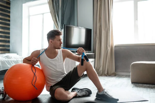 Молодой человек с хорошей фигурой занимается спортом в квартире. Серьезный сконцентрированный парень склоняется к большому красному мячу. Он держит голубую скакалку вокруг шеи . — стоковое фото