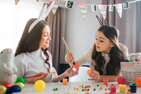 Mutter und Tochter sitzen am Tisch und bemalen Eier. Sie bereiten sich auf Ostern vor. Mädchen halten Pinsel. Mutter und Kind tragen Hasenohren. — Stockfoto
