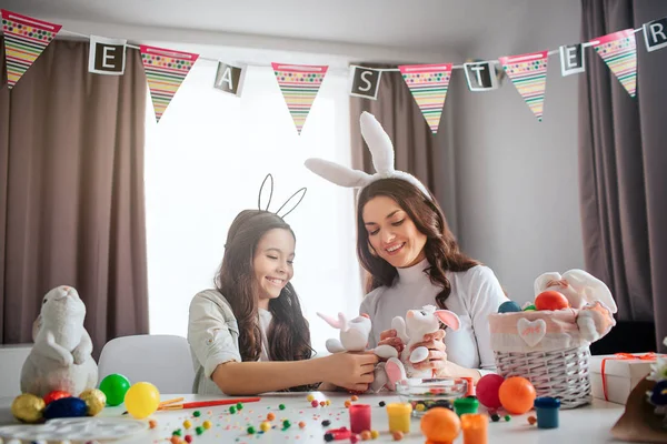 Schöne Mutter und Mädchen bereiten sich auf Ostern vor. Sie spielen zusammen mit weißen Hasenspielzeugen auf dem Tisch. Familie zusammen Spaß haben. Dekoration und Farbe auf dem Tisch. — Stockfoto