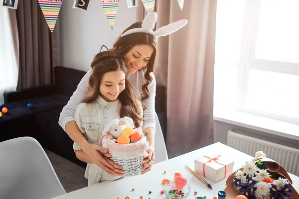 Gut gelaunt stehen Mutter und Tochter am Tisch und bereiten sich im Zimmer auf Ostern vor. Sie halten Korb mit Eiern und Süßigkeiten zusammen. Die Leute tragen Hasenohren auf dem Kopf. Tageslicht. — Stockfoto