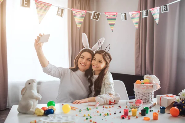 Junge Mutter und Tochter bereiten sich auf Ostern vor. Sie sitzen im Zimmer und machen ein Selfie mit der Handykamera. Dekoration und bunte Eier mit Bemalung auf dem Tisch. Osterhasen. — Stockfoto