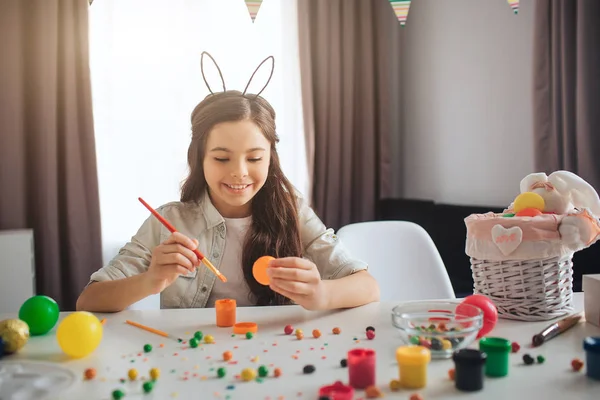 Szczęśliwy nastolatek pozytywne, przygotowanie do Wielkanocy. Ona sama siedzieć w tabeli i malowania jaj. Dziewczyna uśmiech. Nosić uszy królika. Dekoracja i kolorowe cukierki na stole. — Zdjęcie stockowe