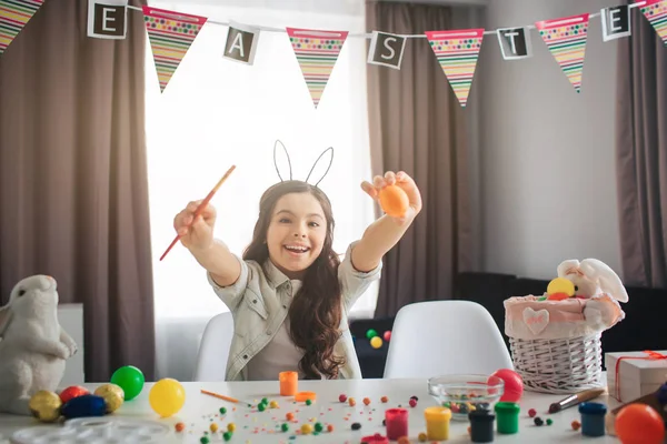 Glückliche positive Mädchen bereiten sich allein auf Ostern vor. Sie sitzt am Tisch im Zimmer und bemalt Eier. Mädchen halten Pinsel und Ei. Lächeln. schön. — Stockfoto