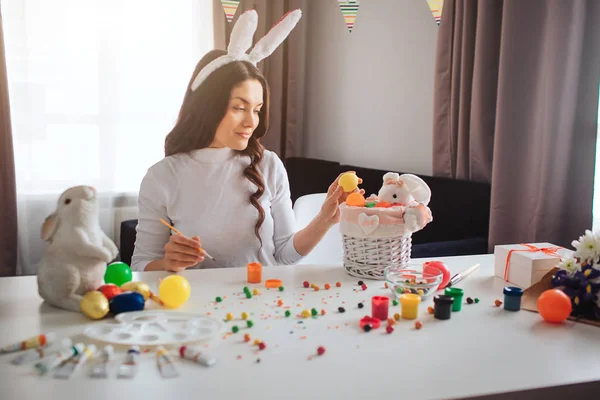 Piękny piękny młoda kobieta przygotować na Wielkanoc. Ona usiąść przy stole i przytrzymaj kolorowy jaj. Model umieścić w kosz z zabawka królik. Dekoracje i słodycze na stole. — Zdjęcie stockowe
