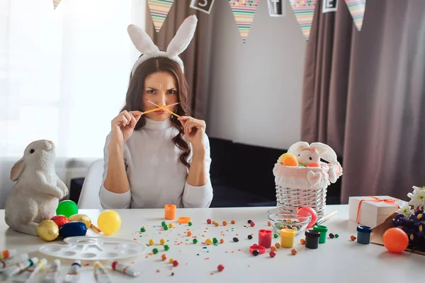 Młoda kobieta przygotować na Wielkanoc sama śmieszne zdenerwowany. Ona siedzieć przy stole w pokoju i przytrzymaj szczotki skrzyżowane. Model spojrzenie na aparat. Ozdoba z farby i słodycze na stole. — Zdjęcie stockowe