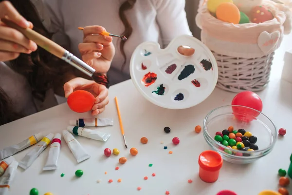 母女俩为复活节做准备。他们拿着刷子, 给鸡蛋涂上一些油漆颜色。碗里的糖果。带玩具和鸡蛋的篮子。颜色的托盘。剪切视图. — 图库照片