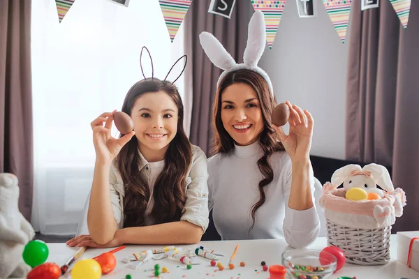 Nette junge Mutter und Tochter bereiten sich auf Ostern im Zimmer vor. Sie halten bunte Eier in den Händen und posieren vor der Kamera. Die Menschen lächeln. Dekoration mit Korb und Farbe auf dem Tisch. — Stockfoto