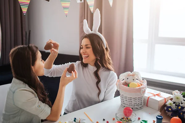 Gut gelaunte Mutter und Tochter bereiten sich auf Ostern vor. Sie halten Schokoladeneier in der Hand und lächeln. Tischdekoration. Modell trägt weiße Kaninchenohren. — Stockfoto