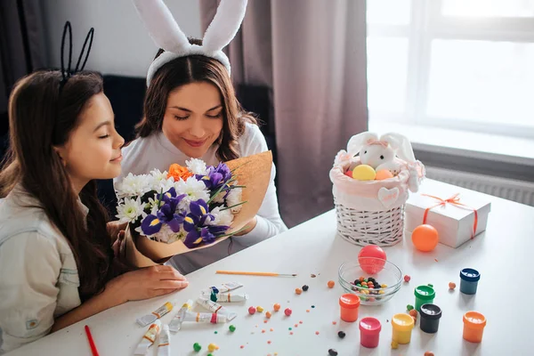 Junge Frau und ihre Tochter bereiten sich auf Ostern vor. sie riechen gemeinsam Blumen. Farbe mit Süßigkeiten und Dekoration auf dem Tisch. Tageslicht. — Stockfoto