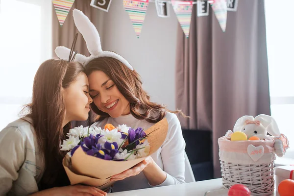 Ładny Mama i córka przygotować razem na Wielkanoc w pokoju. One siedzieć i przytulić się. Młoda kobieta trzymać piękny bukiet kwiatów. Spokojna i szczęśliwa. — Zdjęcie stockowe