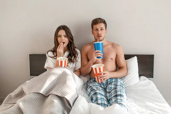 Młoda para w łóżku. Zaintrygowany piękny mężczyzna i kobieta są jedzenie popcorn i oglądania telewizji razem w sypialni — Zdjęcie stockowe