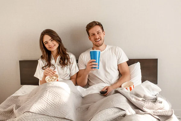 Casal jovem na cama. Sorrindo belo homem e mulher estão comendo pipocas e assistindo TV juntos no quarto — Fotografia de Stock