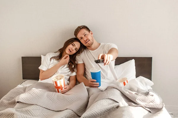 Casal jovem na cama. Sorrindo belo homem e mulher estão segurando controle remoto e comer pipocas enquanto assiste TV — Fotografia de Stock