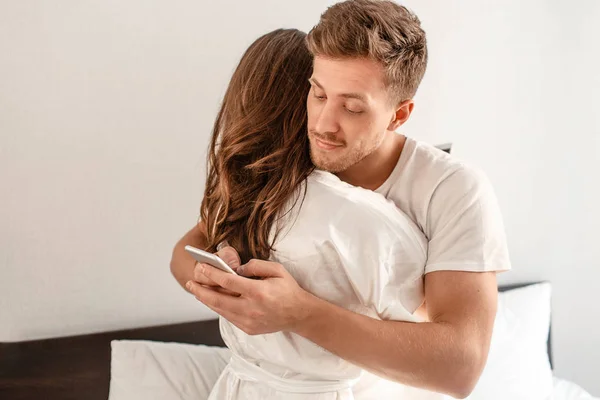 Pareja joven en el dormitorio. Sonreír hombre infiel es engañar y enviar mensajes de texto amante en el teléfono mientras abraza a su novia — Foto de Stock
