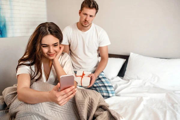 Pareja joven en la cama. Sonriendo mujer indiferente con su teléfono está ignorando a su hombre infeliz con un regalo — Foto de Stock