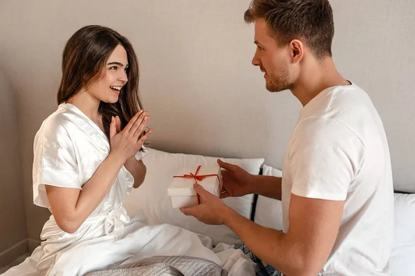 Casal jovem na cama. Um homem atraente está dando o presente para sua namorada feliz, romance em um relacionamento — Fotografia de Stock