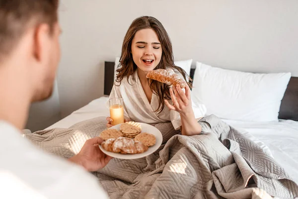 Casal jovem com delicioso café da manhã na cama. Mulher bonita está comendo croissant fresco com suco — Fotografia de Stock