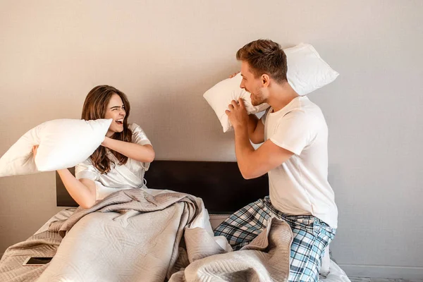 Młoda para w łóżku. Uśmiechający się zadowolony, mężczyzna i kobieta w piżamy są oszukiwanie i walki z poduszkami, dzień dobry — Zdjęcie stockowe